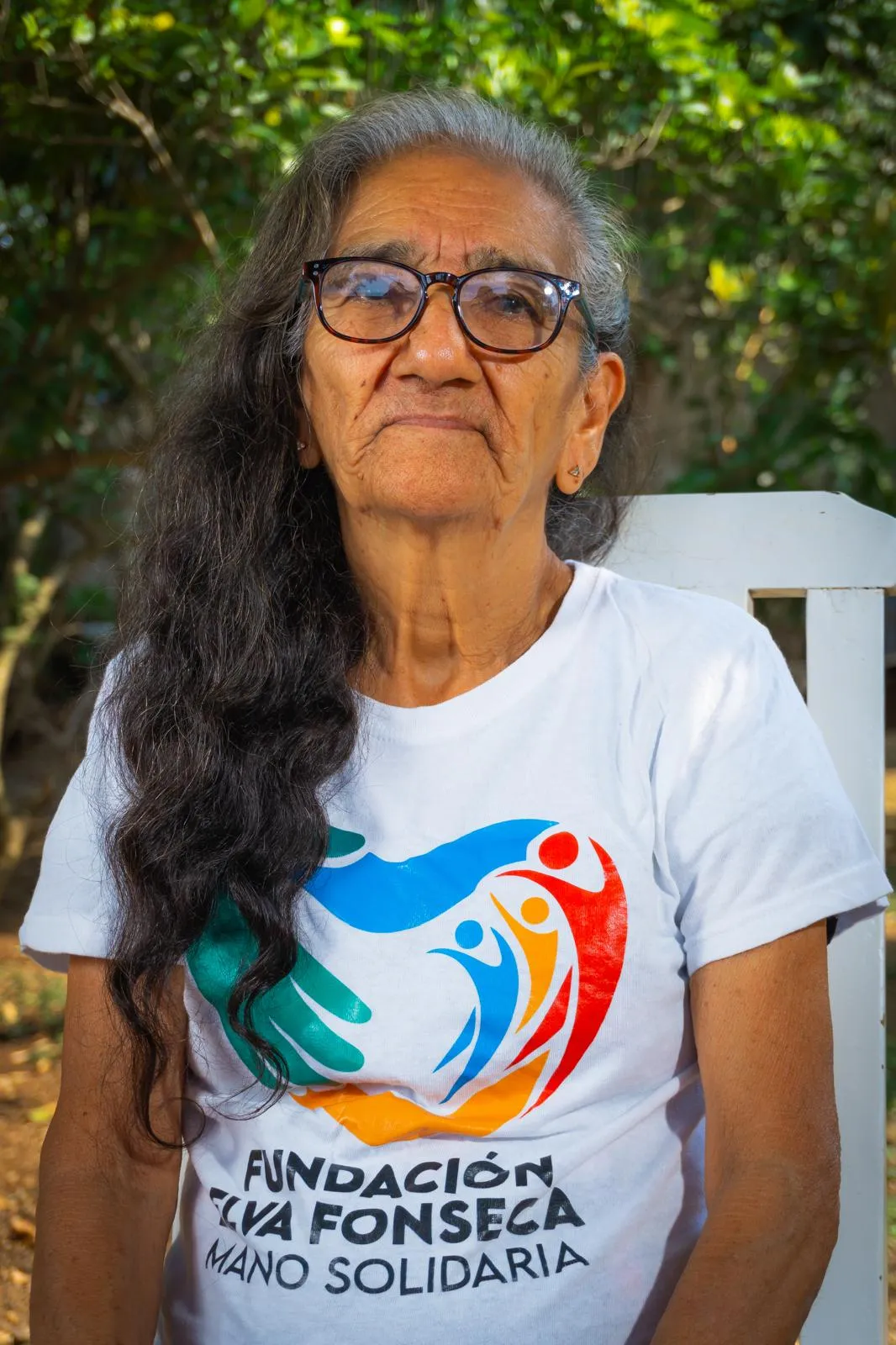 An elderly woman wearing glasses and a Fundación Elva Fonseca t-shirt, outdoors